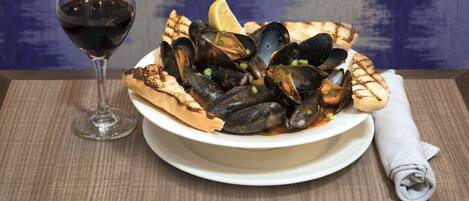 A plate of mussels with grilled bread and lemon wedges, accompanied by a glass of red wine on a table with a napkin.