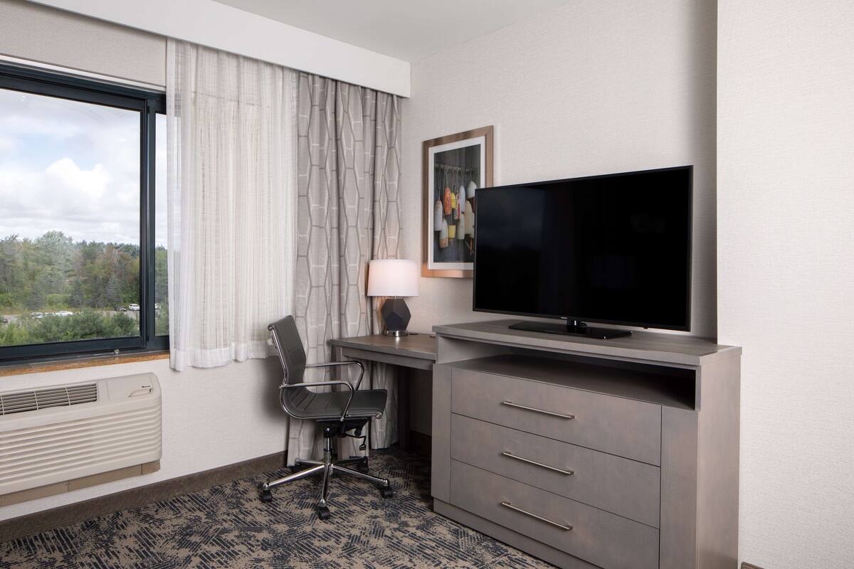Une chambre d’hôtel dotée d’une grande fenêtre, d’un bureau avec un téléviseur à écran plat, d’une chaise et d’une lampe.