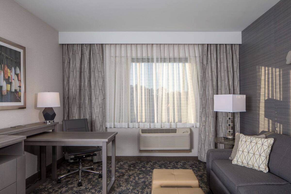 Une chambre d’hôtel équipée d’un bureau, d’une chaise, d’un canapé et d’une grande fenêtre avec des rideaux.