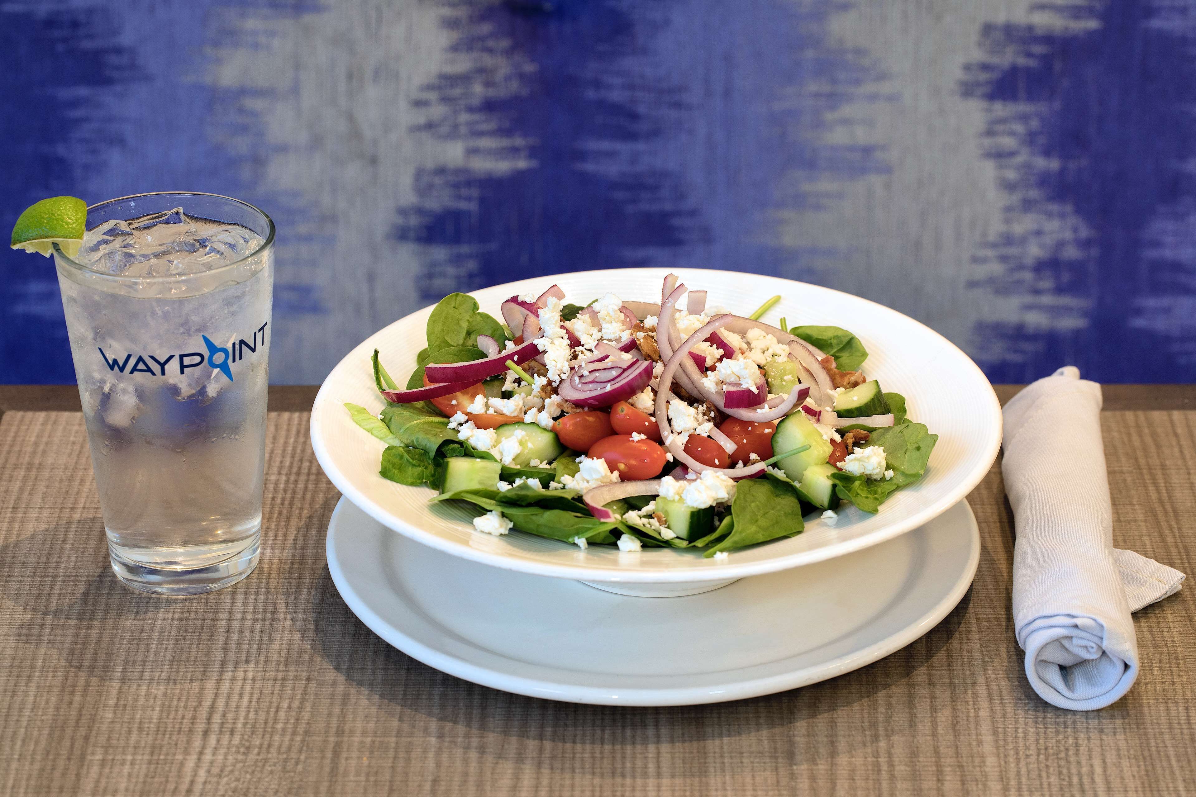 Um prato branco com salada, um copo de água e uma toalha de tecido dobrada sobre uma mesa.