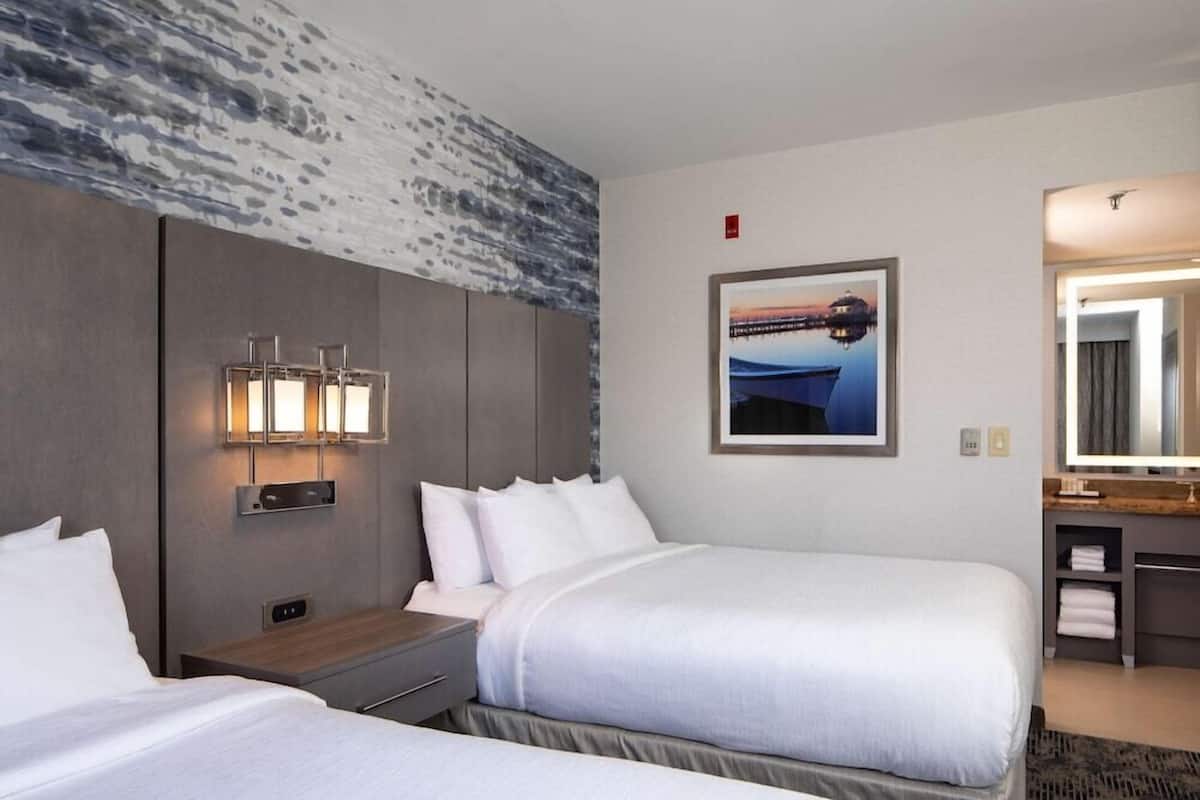 Une chambre d’hôtel avec deux lits, un grand luminaire fixé au mur, un tableau représentant un coucher de soleil sur l’eau et un miroir au-dessus de la coiffeuse.