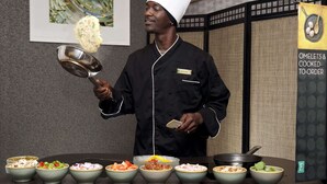 A chef in a black uniform and white hat, holding a ladle and a bowl, with various dishes in front of him. - Embassy Suites by Hilton Portland Maine (Portland)