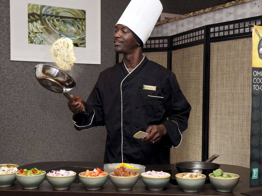 A chef in a black uniform and white hat, holding a ladle and a bowl, with various dishes in front of him.