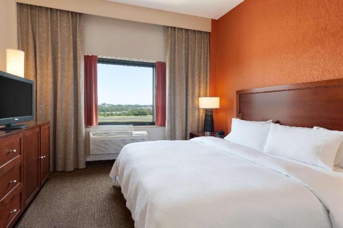 Una habitación de hotel con una cama, un televisor sobre un mueble de madera, una ventana con cortinas y una mesita de noche con lámpara.