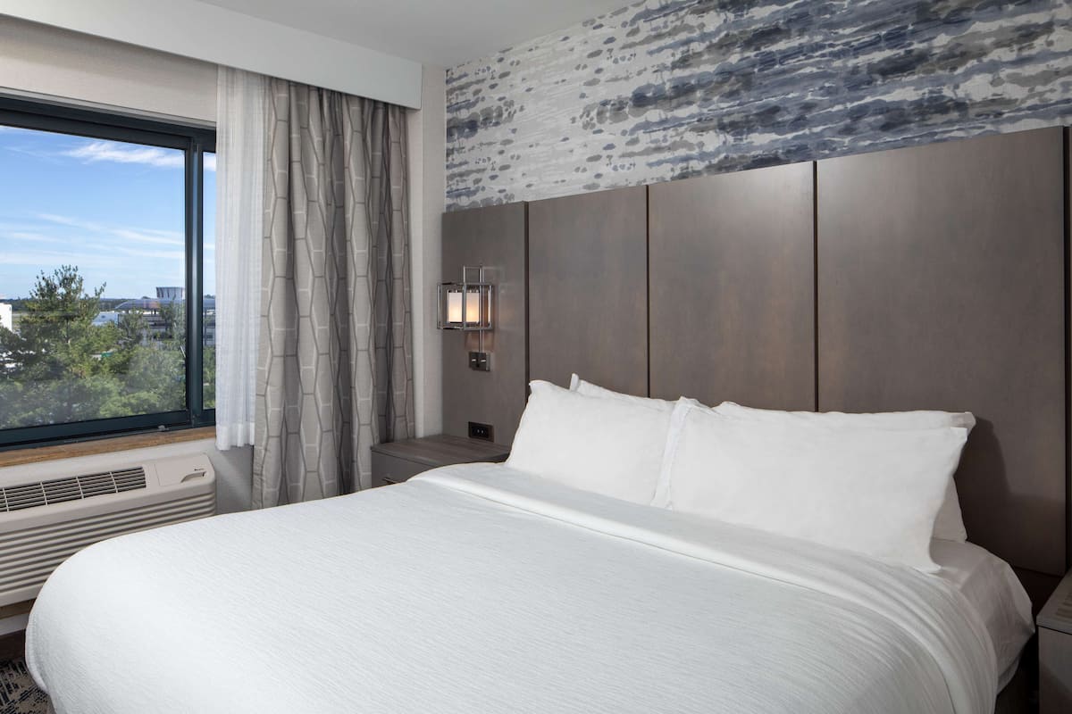 Une chambre d’hôtel avec un grand lit, une table de chevet, une lampe fixée au mur et une fenêtre avec des rideaux.