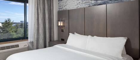 Habitación de hotel con una cama grande, una mesita de noche, una lámpara de pared y una ventana con cortinas.