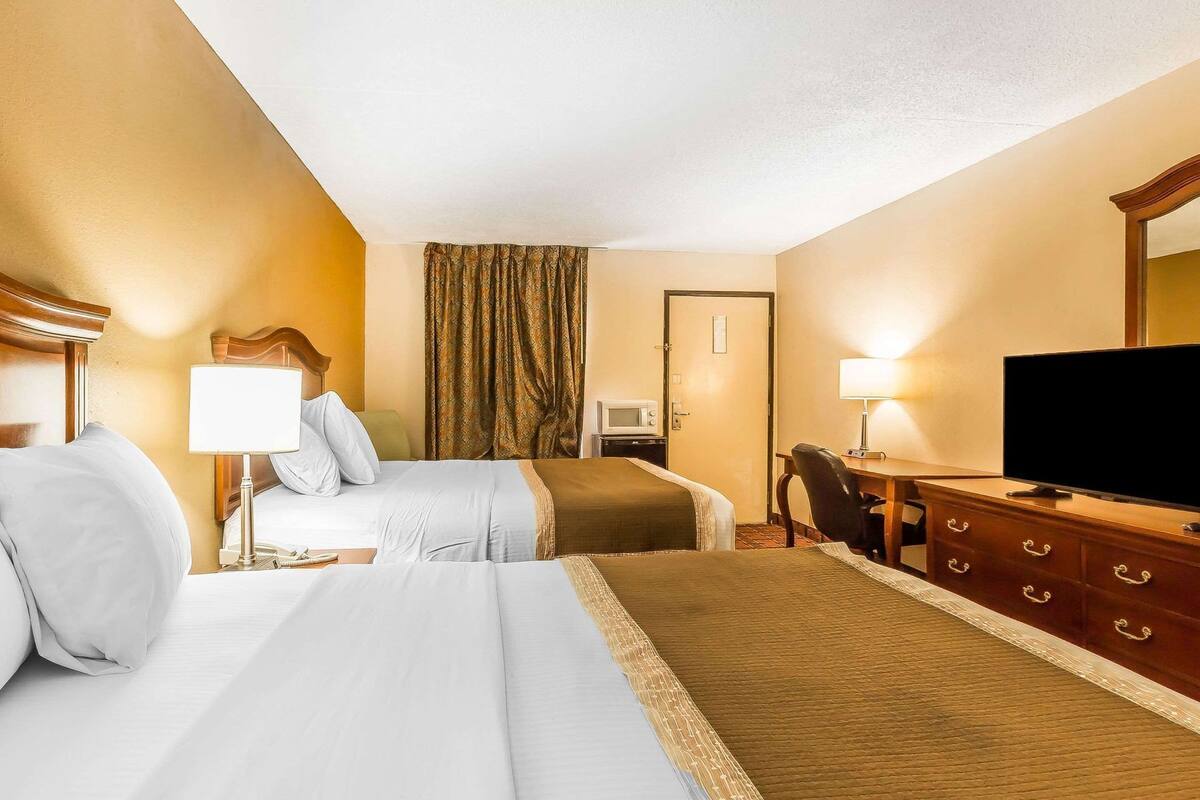 A hotel room with two beds, a television, a desk with drawers, a chair, a microwave, and a window with curtains.