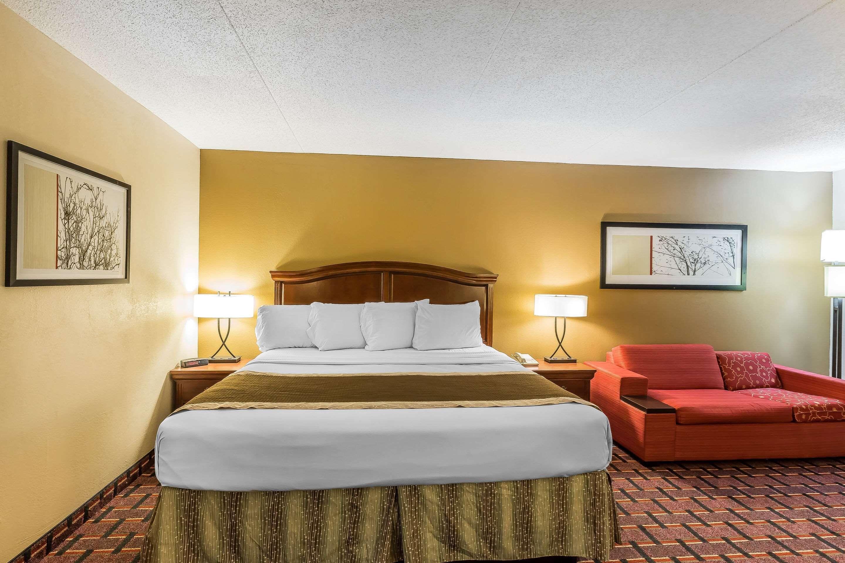 Habitación de hotel con una cama grande, un sillón rojo, dos lámparas de noche y obras de arte en las paredes.