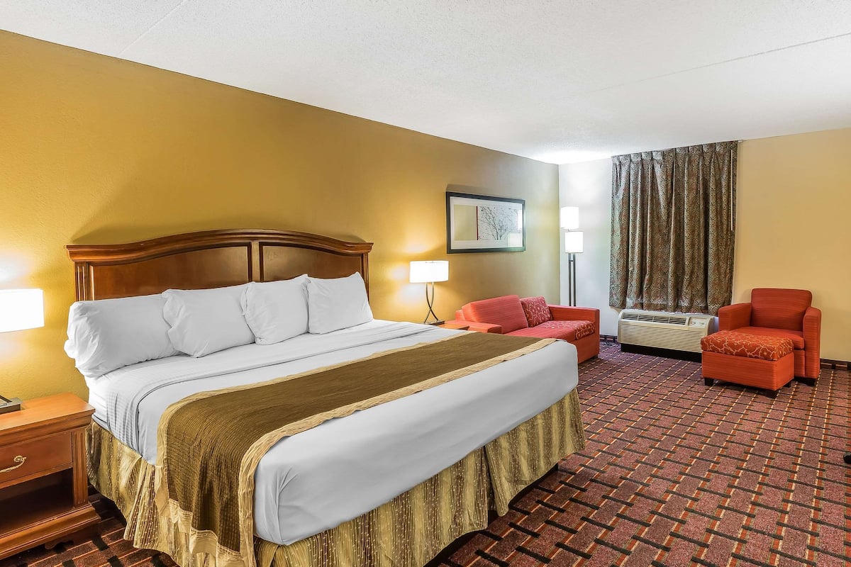 Habitación de hotel con una cama grande, dos sillas, una mesita de noche, una lámpara y una ventana con cortinas.