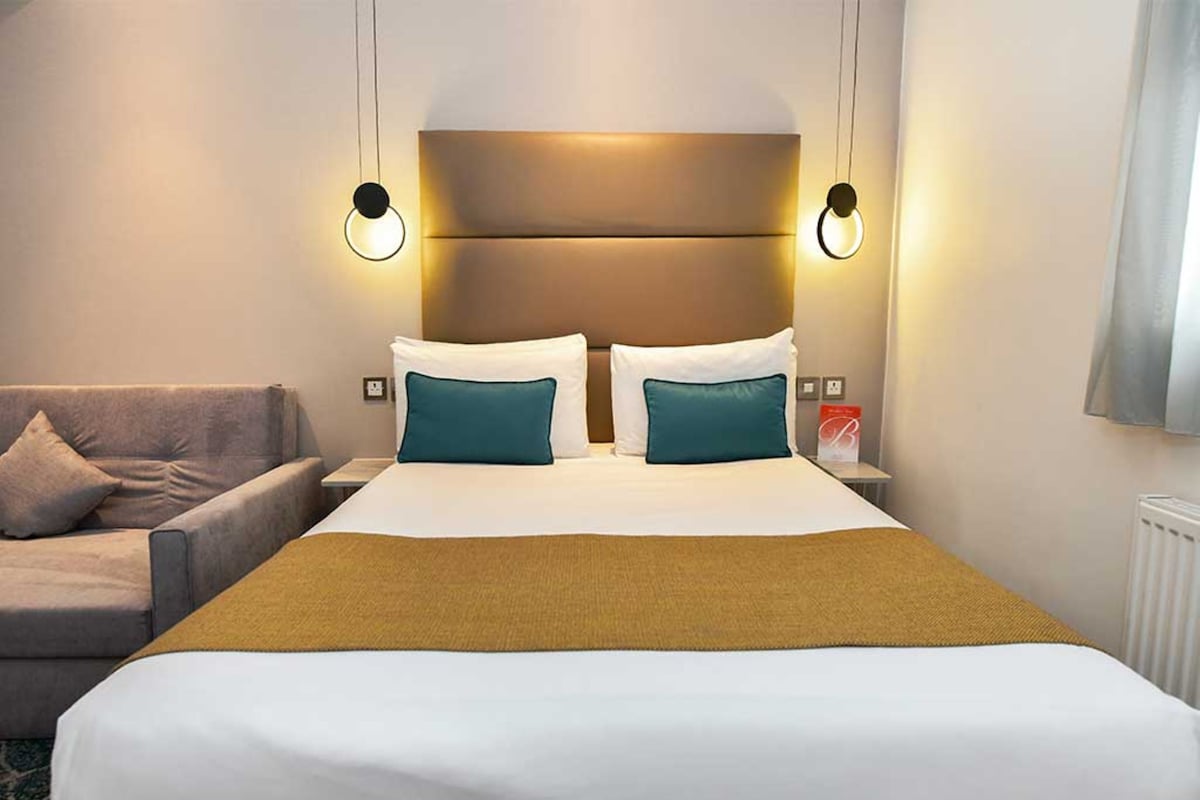 A hotel room with a large bed, two bedside lamps, a nightstand, a sofa, and a window.