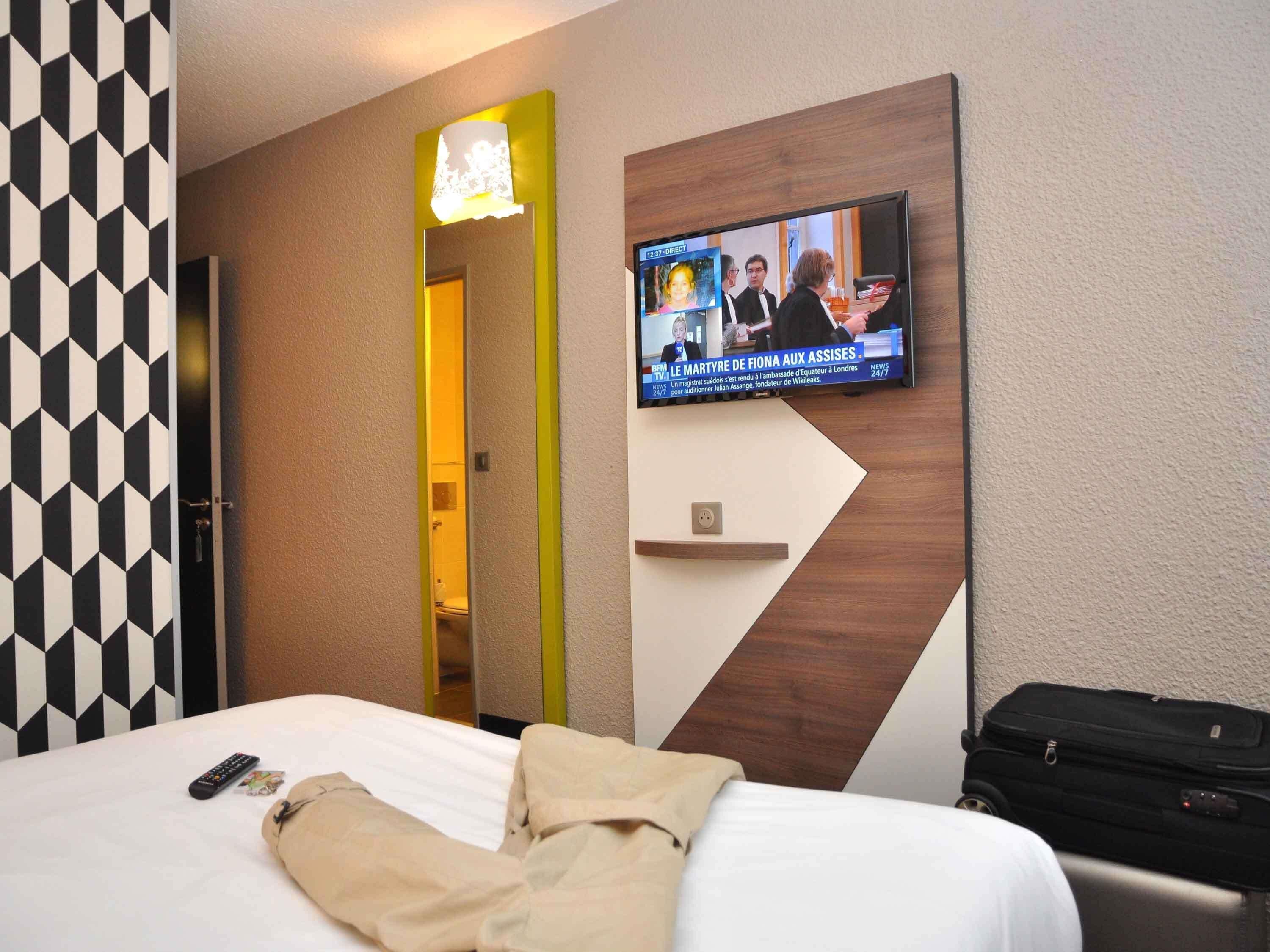 A hotel room with a bed, a television mounted on the wall, a mirror, and a suitcase.