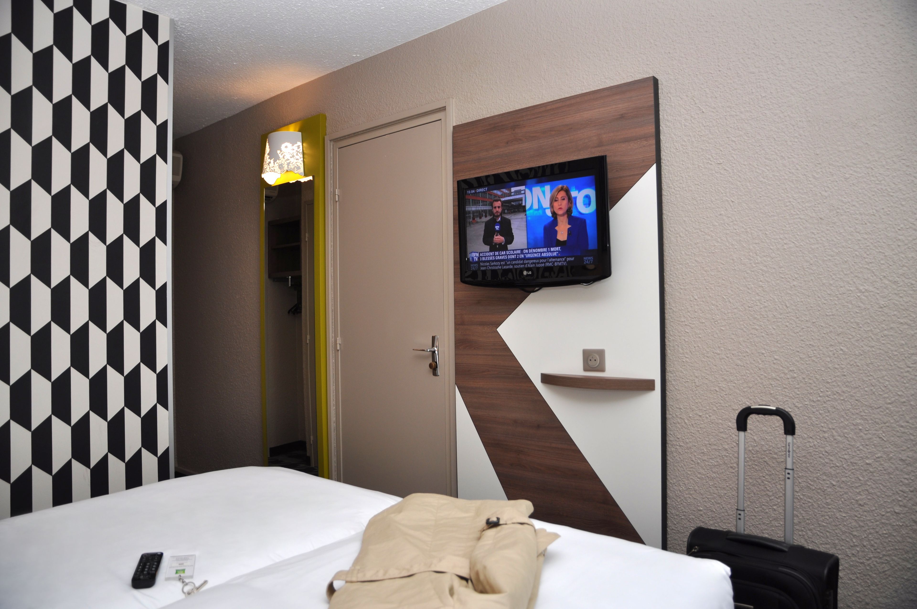 A hotel room with a bed, a TV mounted on the wall, a suitcase, and a door leading to another room.