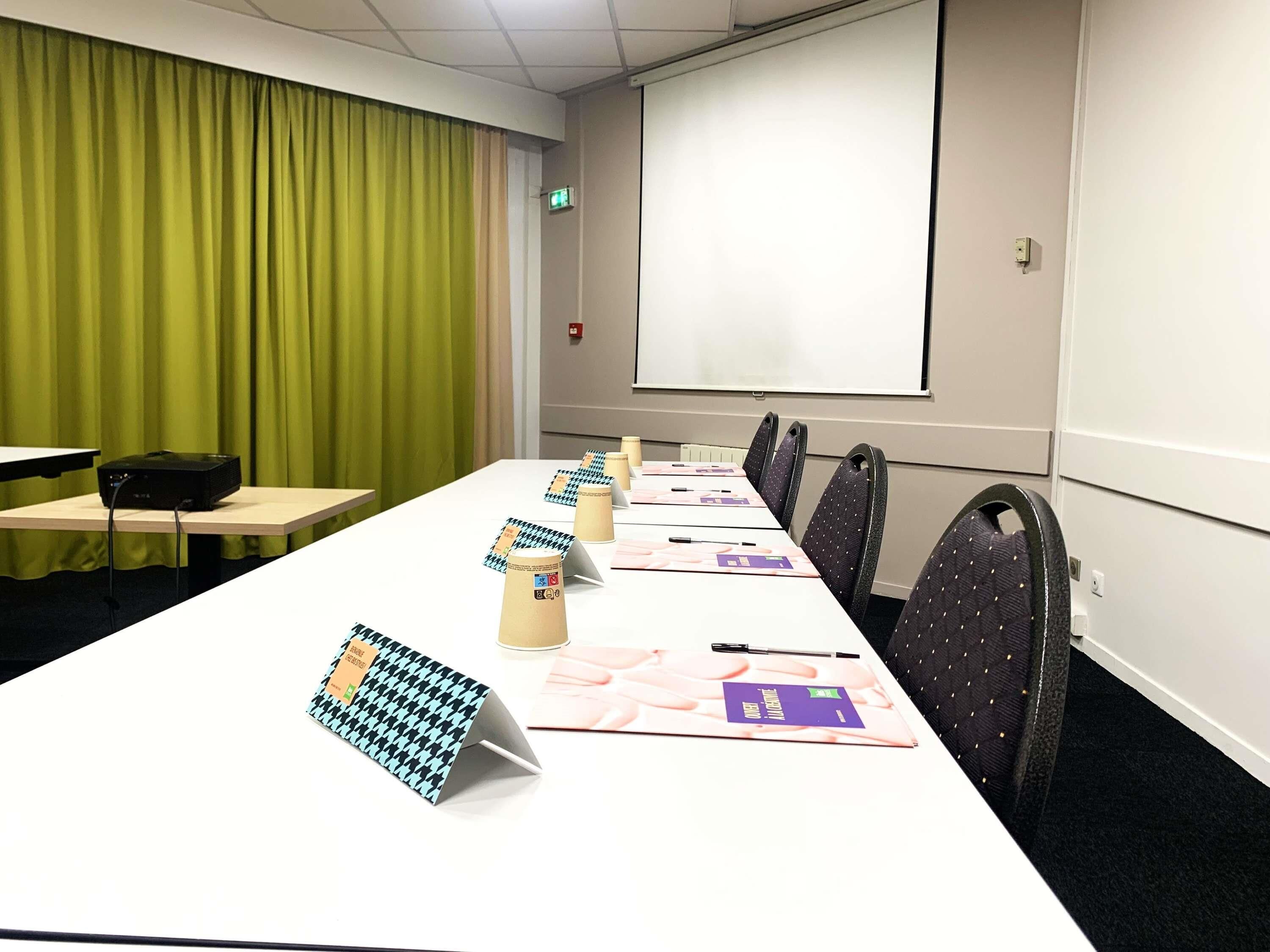 A conference room with a long table, chairs, a projector screen, and a green curtain.