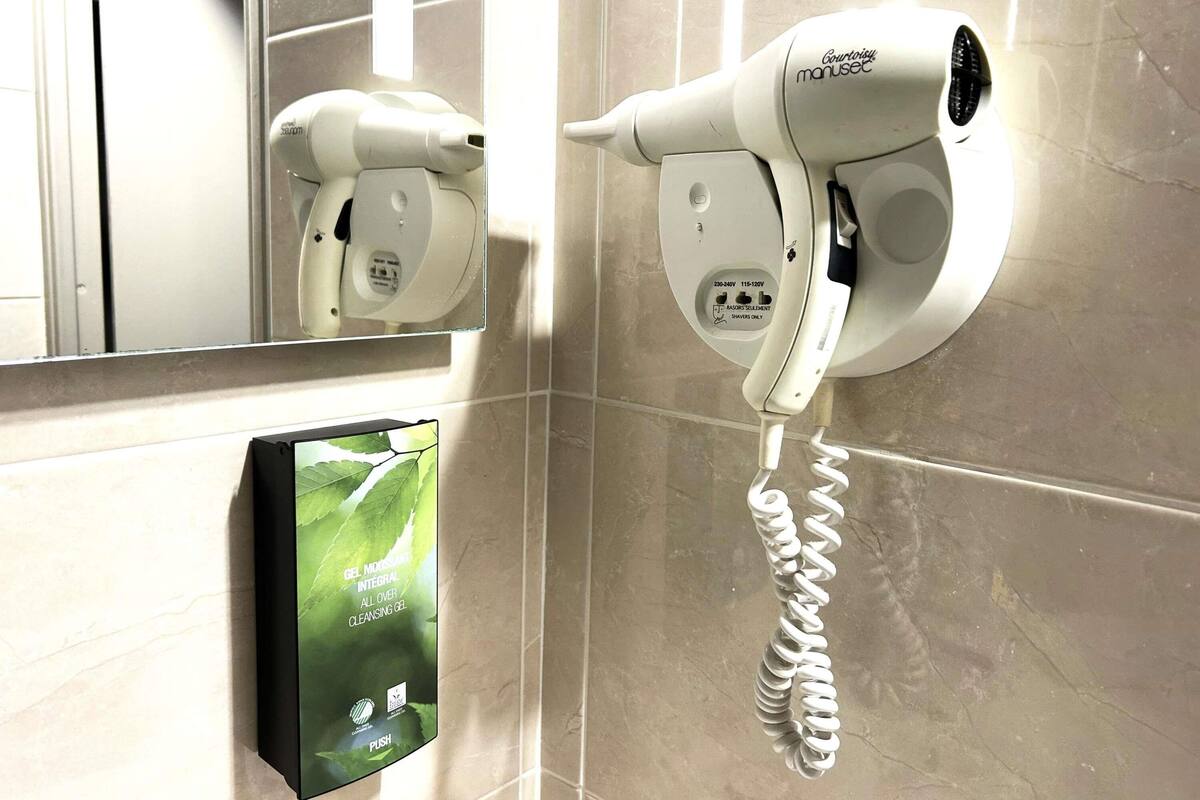 A hairdryer mounted on a wall with a mirror above it and a push-button phone with a charging cable.