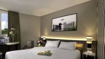 A hotel room with a neatly made bed, a bedside table with a lamp, a desk with a laptop, and a window with curtains.