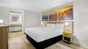 Een moderne hotelkamer met een groot bed, een houten nachtkastje, een spiegel en een schilderij van een zonsondergang aan de muur.
