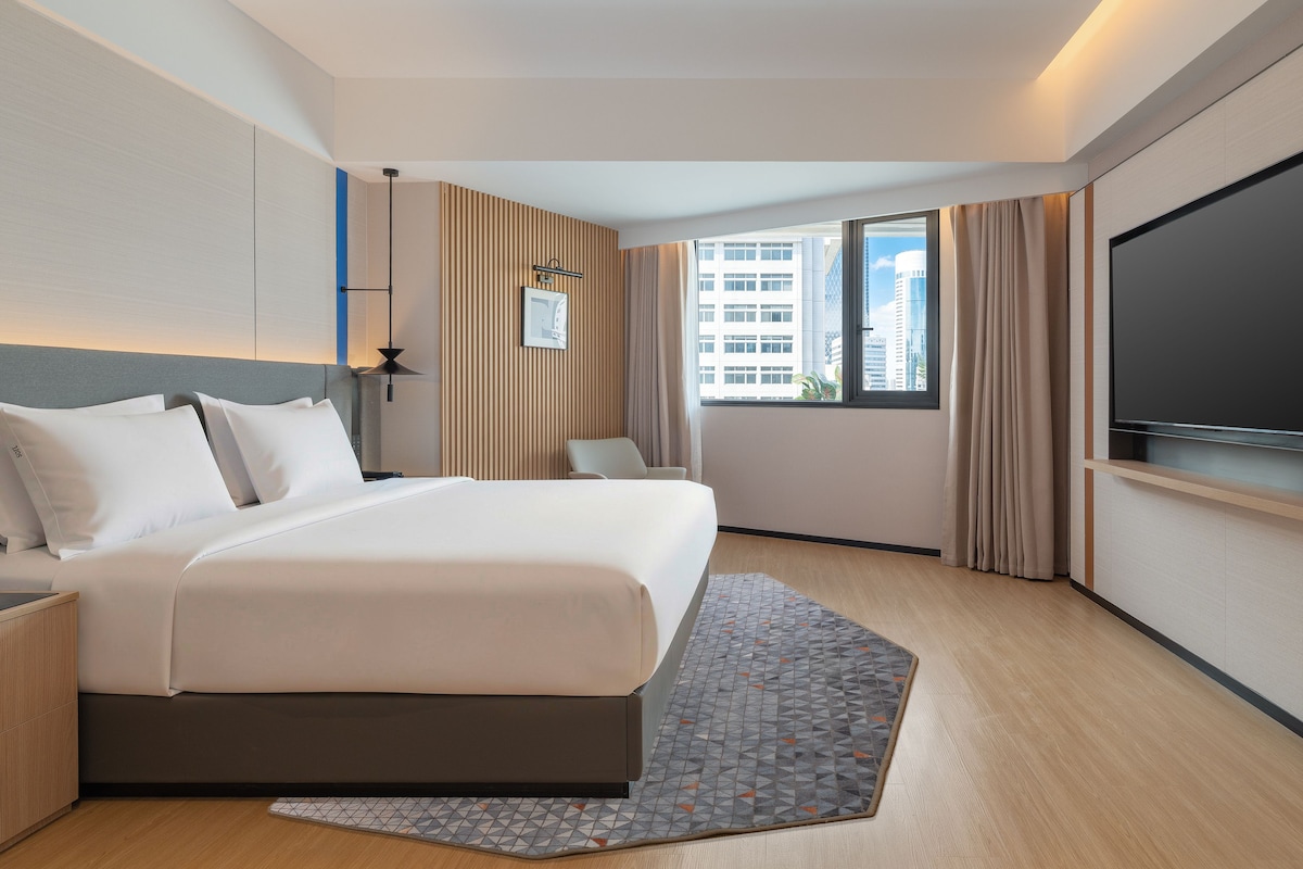 A modern hotel room with a large bed, a flat-screen TV, a desk, and a window with a city view.