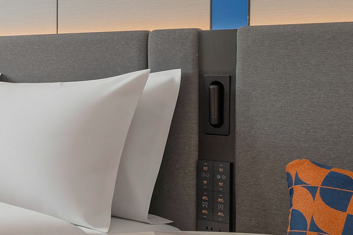 A hotel room with a bed, a control panel, and a patterned throw pillow.