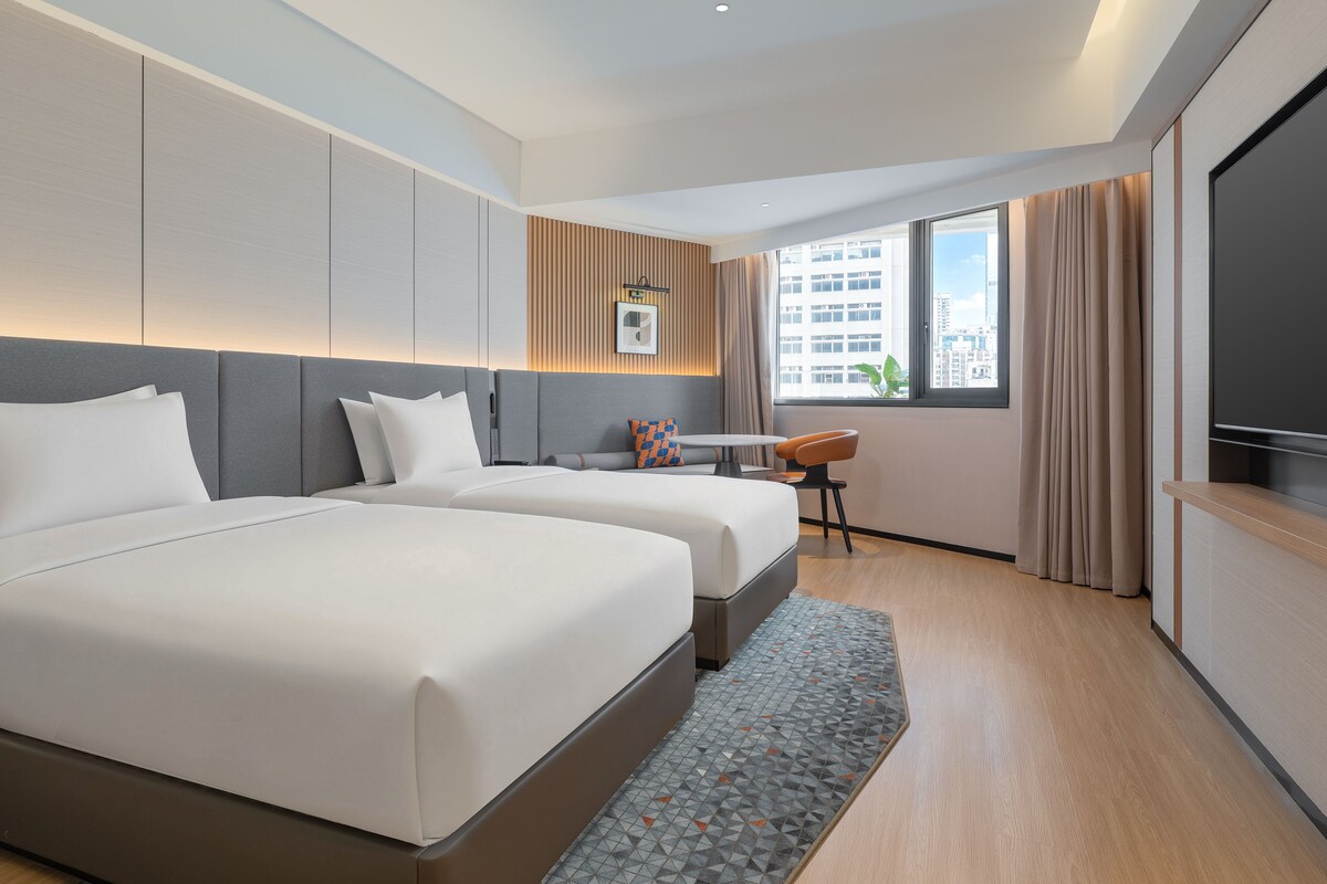 A hotel room with two beds, a desk, a chair, a TV, and a window with a city view.
