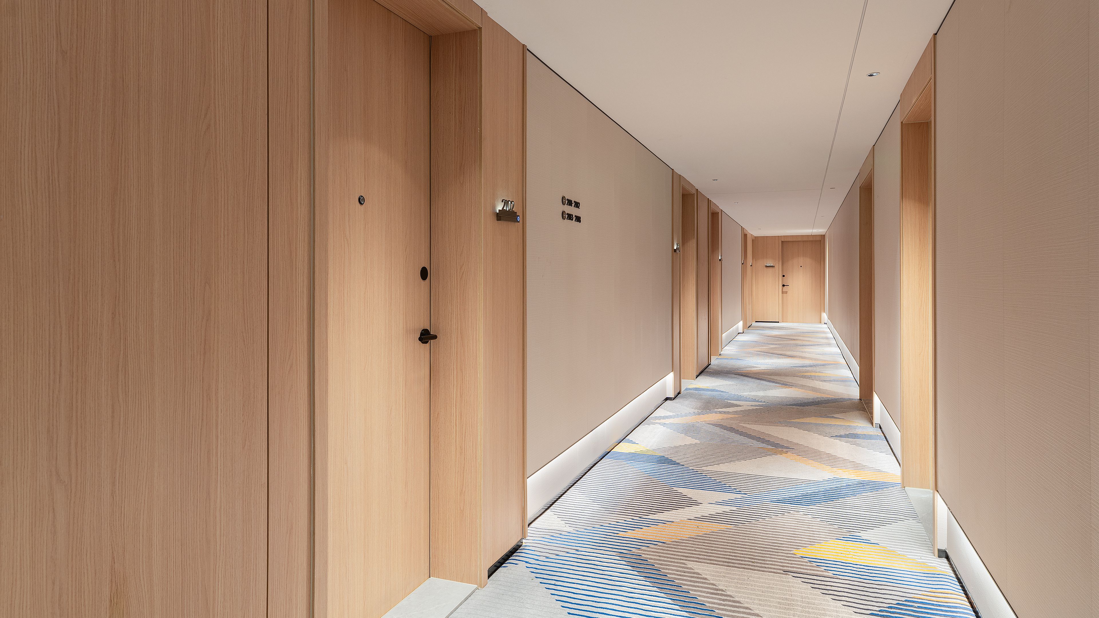 Pasillo de hotel con puertas de madera, alfombra estampada y un cartel que indica la habitación 702.