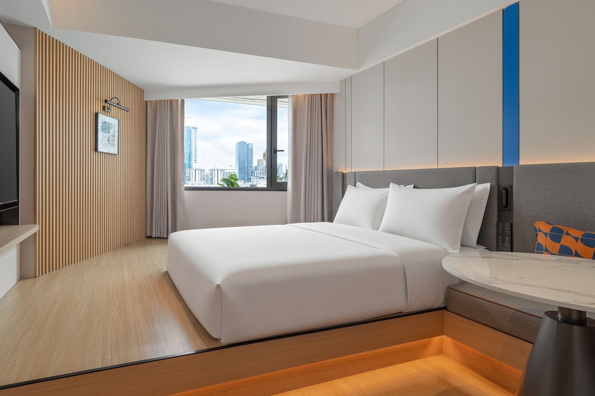 A hotel room with a bed, a desk, a TV, and a window with a city view.