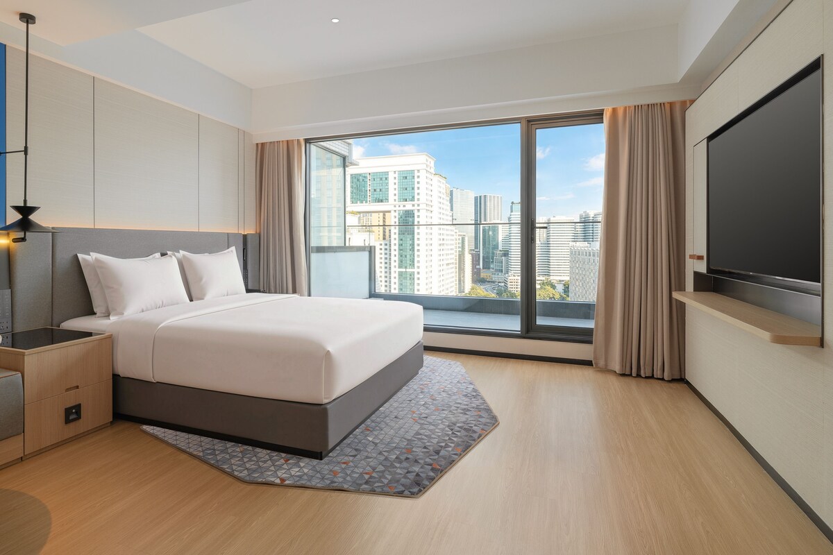 A modern hotel room with a large bed, a desk, a flat-screen TV, and a city view.