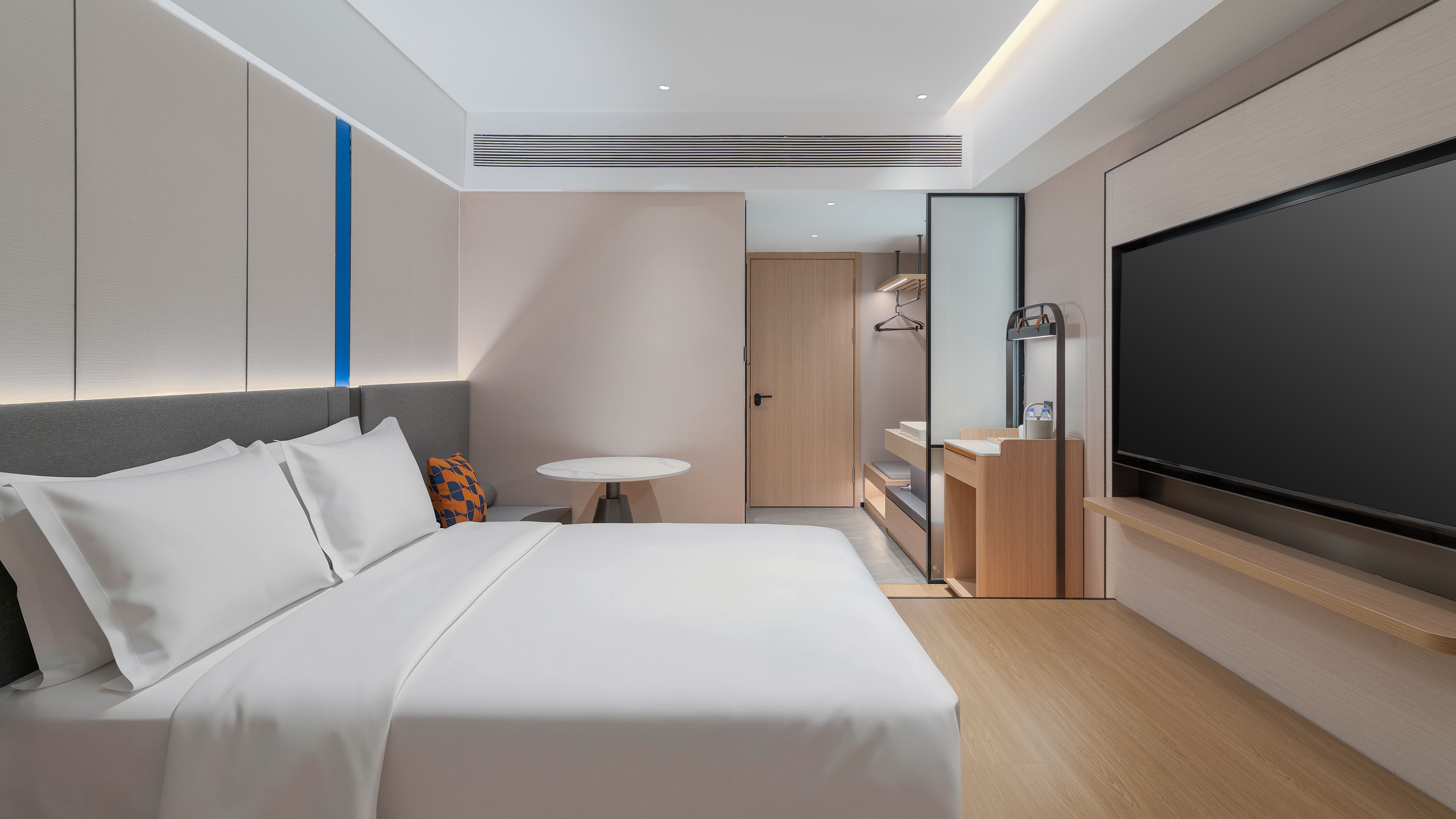 A modern hotel room with a bed, a small round table, a TV, and a bathroom with a mirror and sink.