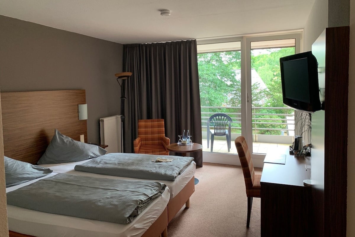 A hotel room with two beds, a desk with a television, a chair, and a view of greenery outside.