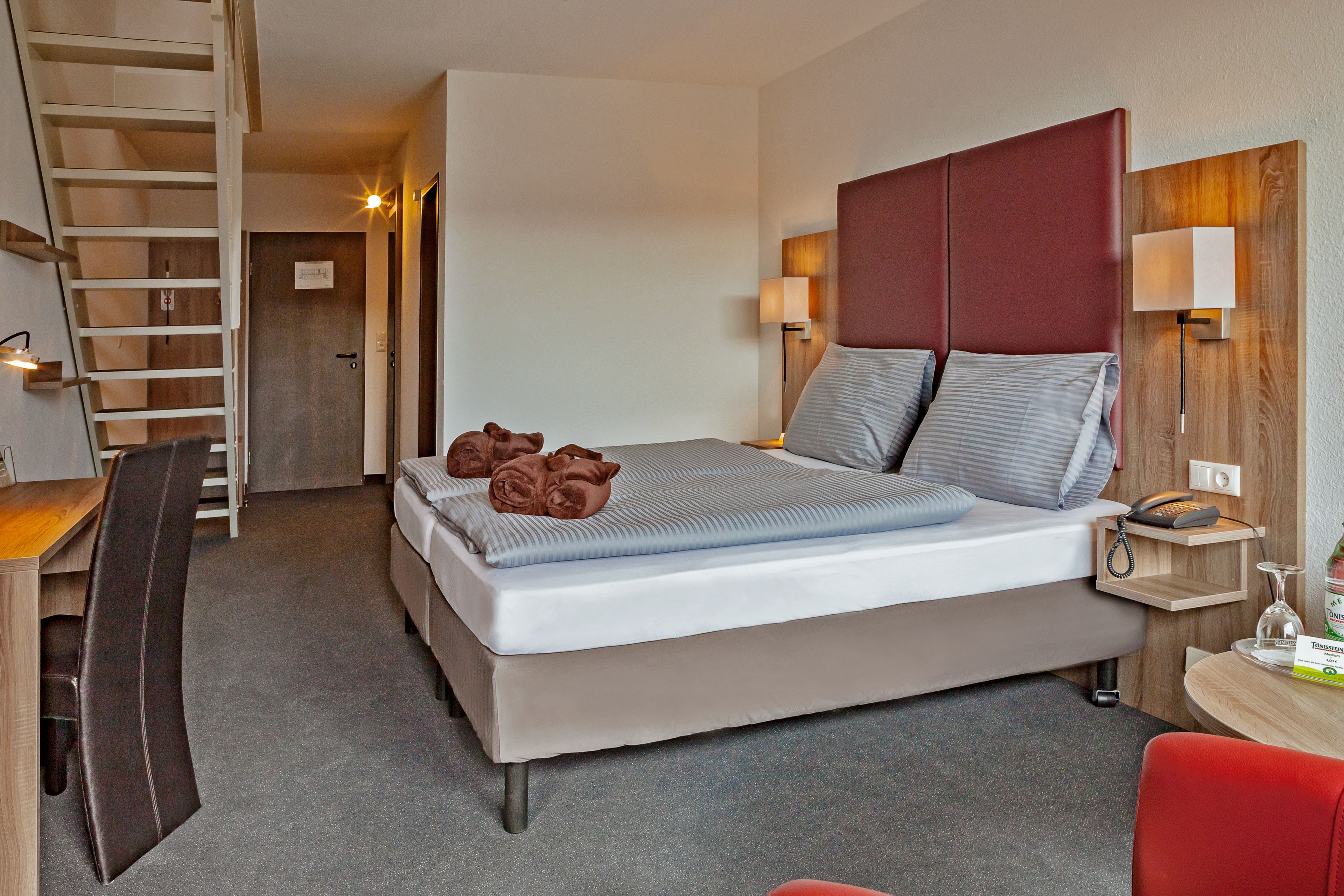 A hotel room with a bed, a desk, a chair, a desk lamp, a bedside table, a phone, and a small table with a bottle and glasses.