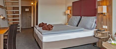 A hotel room with a bed, a desk, a chair, a desk lamp, a bedside table, a phone, and a small table with a bottle and glasses.