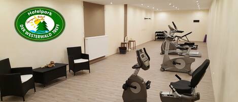 Gimnasio de hotel con máquinas de cardio, un logotipo en la pared y una zona de estar.