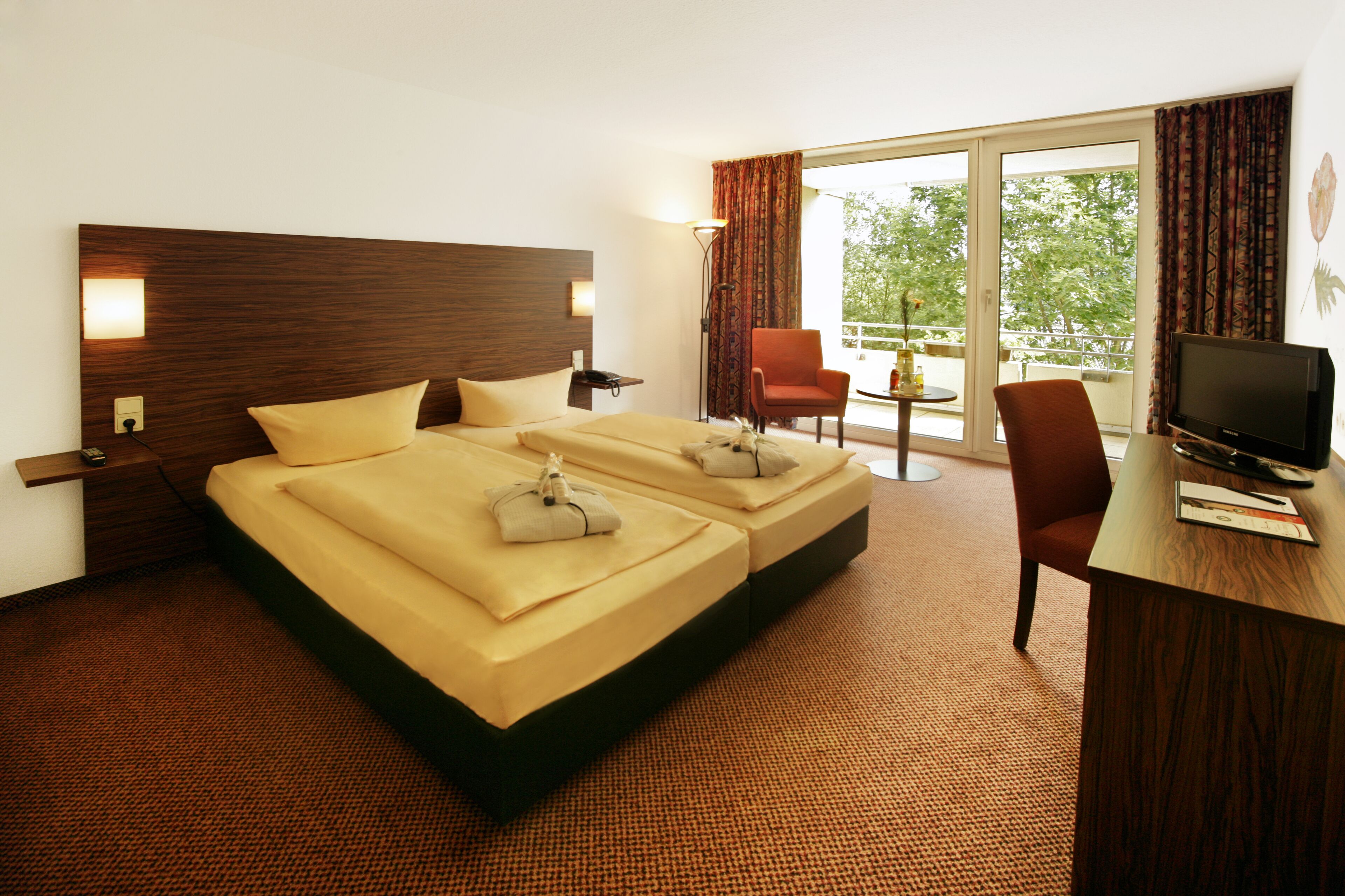 A hotel room with a large bed, a desk with a TV, and a balcony with a chair.