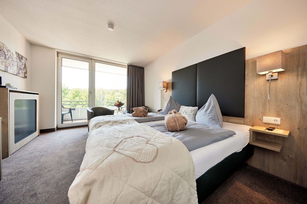 A modern hotel room with a large bed, a desk, a chair, and a balcony with a view.
