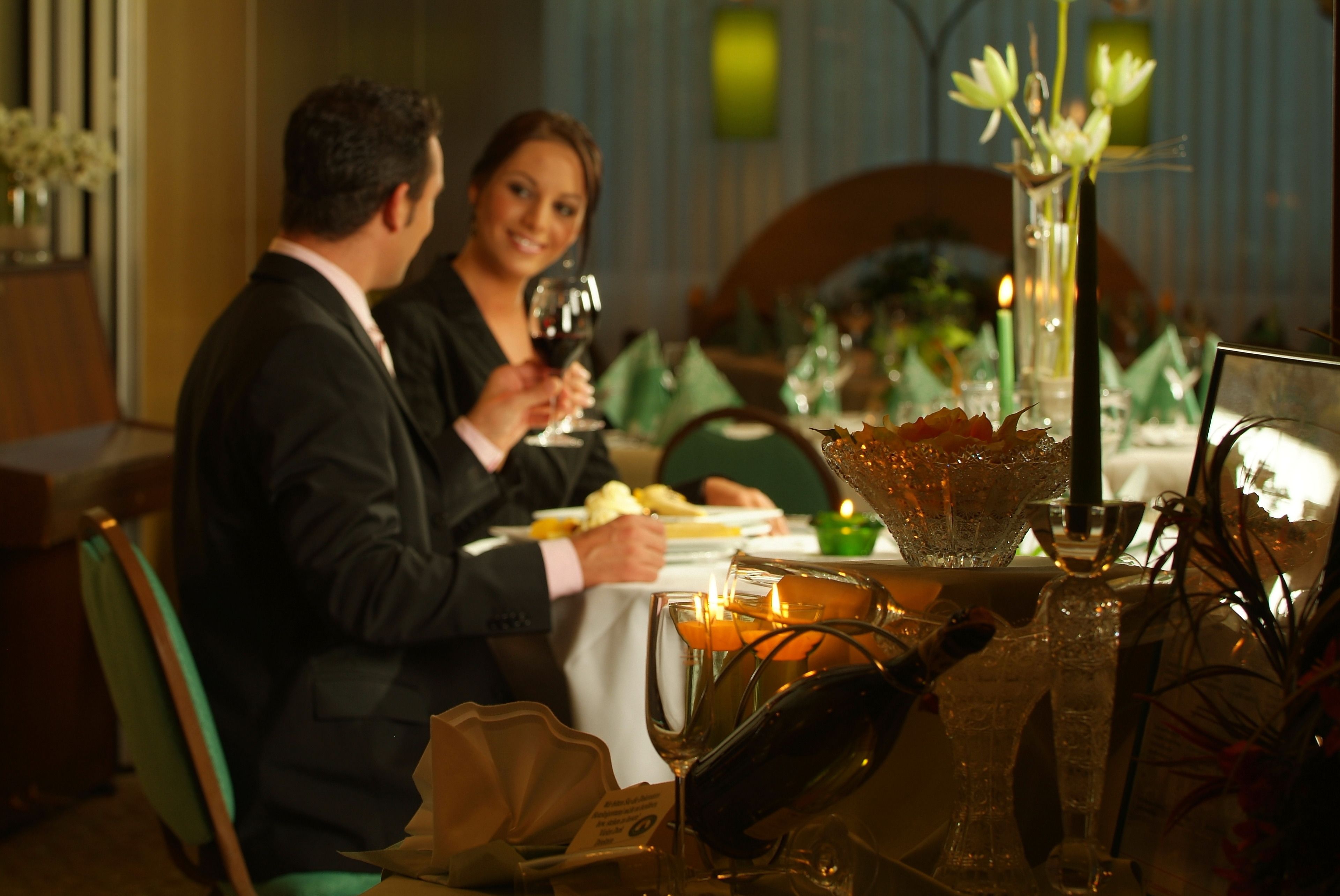 Un couple est assis à une table dans un restaurant, éclairée par des bougies. Un vase rempli de fleurs et un présentoir à menus complètent le décor.