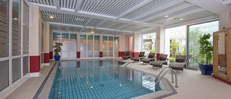 Una piscina interior moderna con agua cristalina, rodeada de sillones de descanso y plantas en macetas.