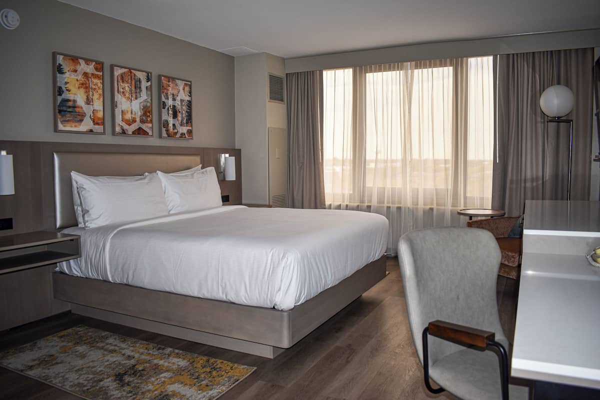A modern hotel room with a large bed, a desk, and a chair. There are three artwork pieces on the wall and a large window with curtains.