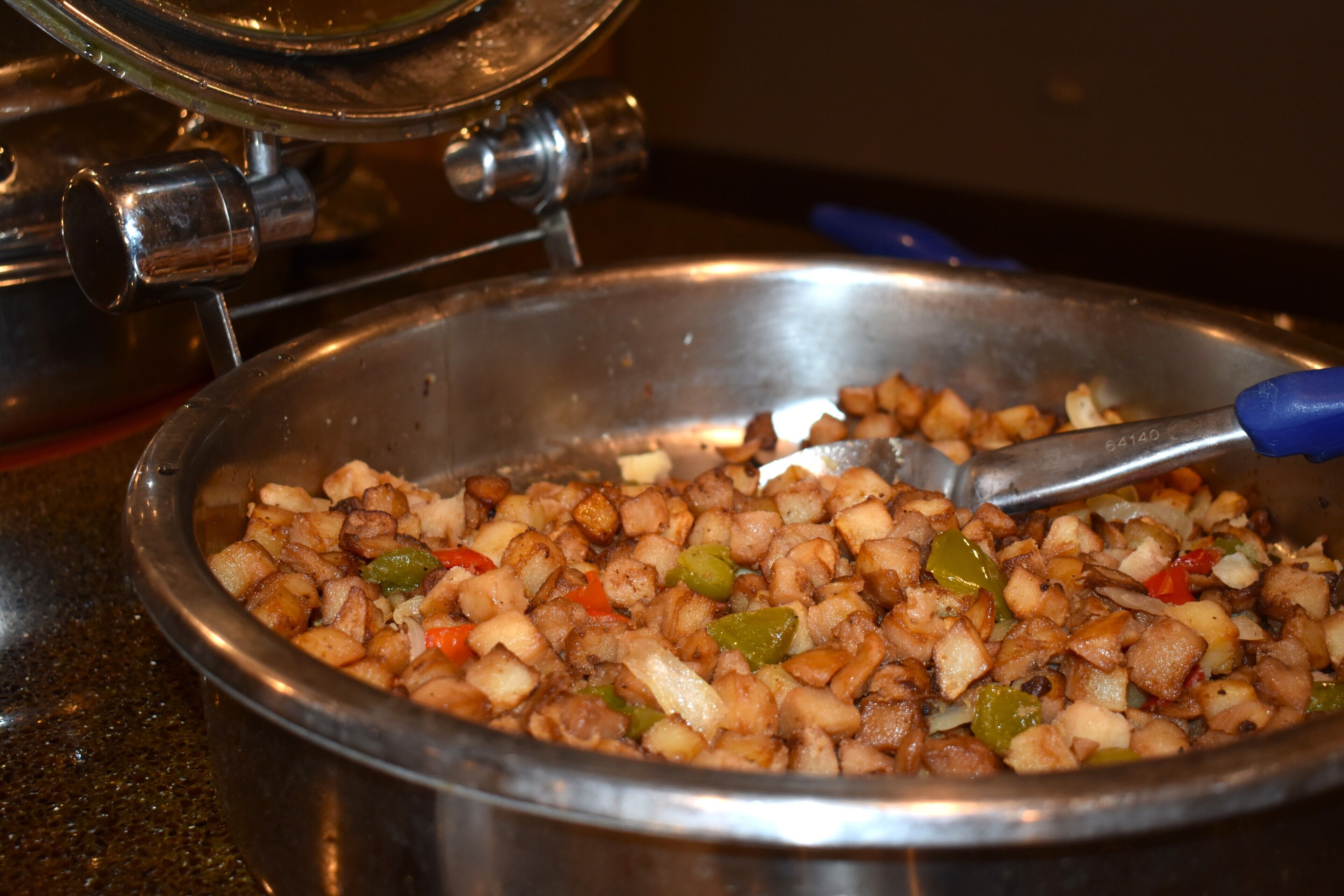 A stainless steel pan filled with diced potatoes, onions, and bell peppers being stirred with a blue-handled utensil.