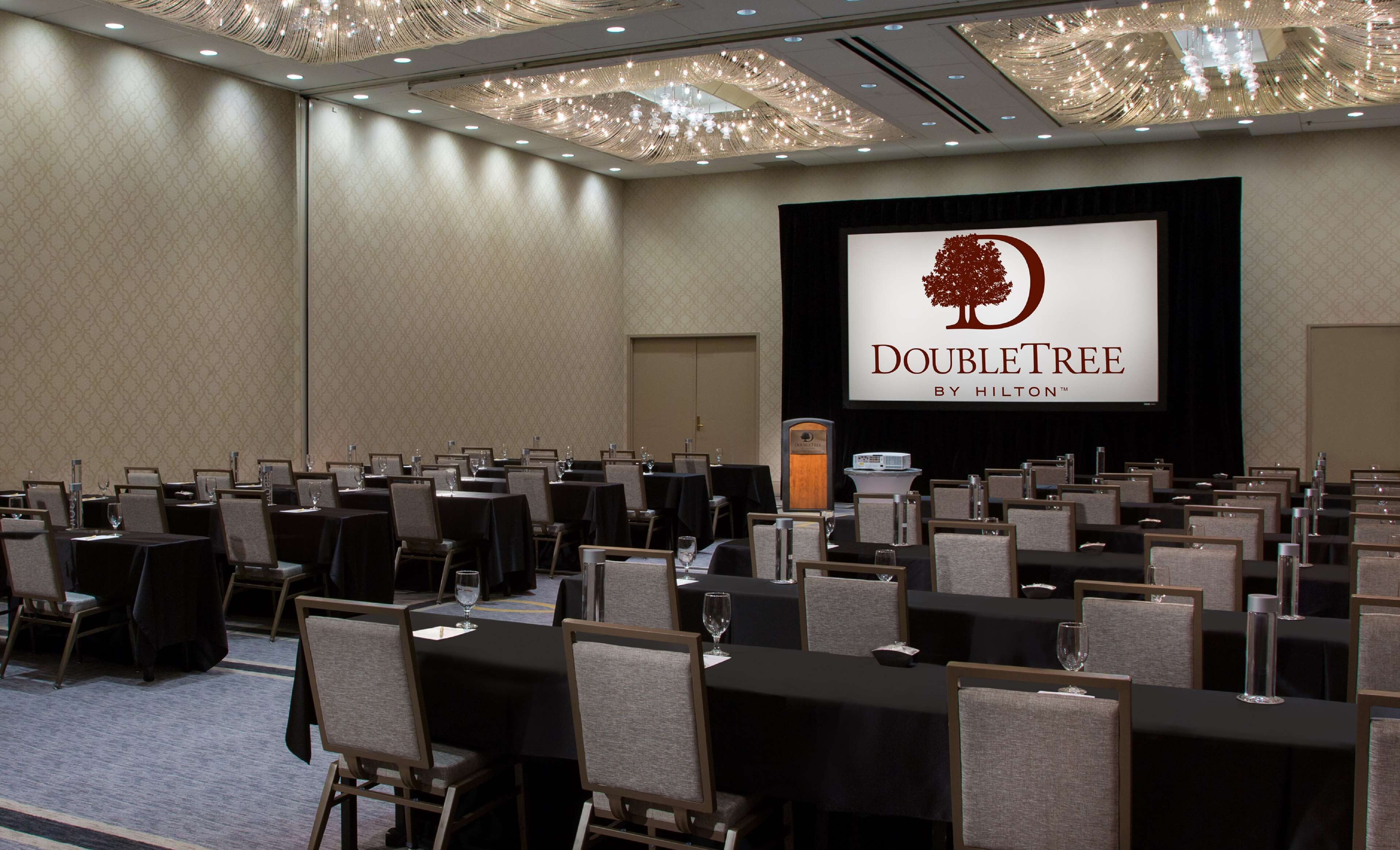 A conference room with rows of tables and chairs, a podium, and a screen displaying the DoubleTree by Hilton logo.