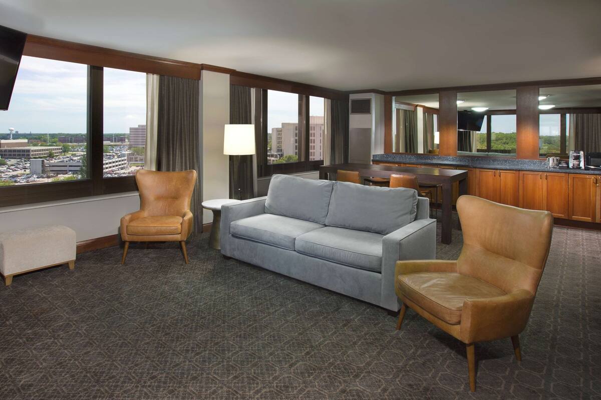 A hotel room with a sofa, two armchairs, a small table, a TV, a kitchen area, and large windows with a city view.