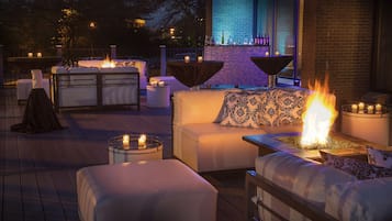 Een terras met witte, beklede zitbanken, ronde tafels met kaarsen en een open haard in het midden.