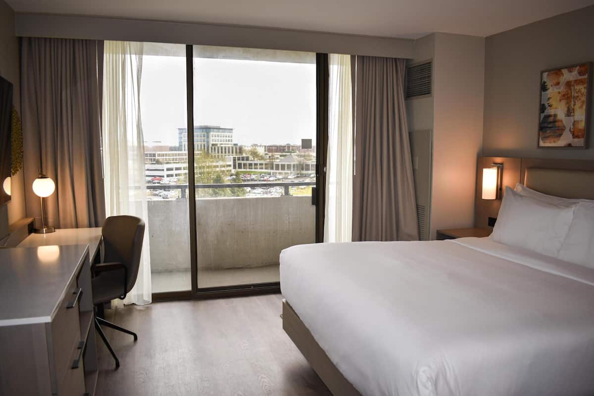 A hotel room with a bed, desk, chair, and a large window with curtains.