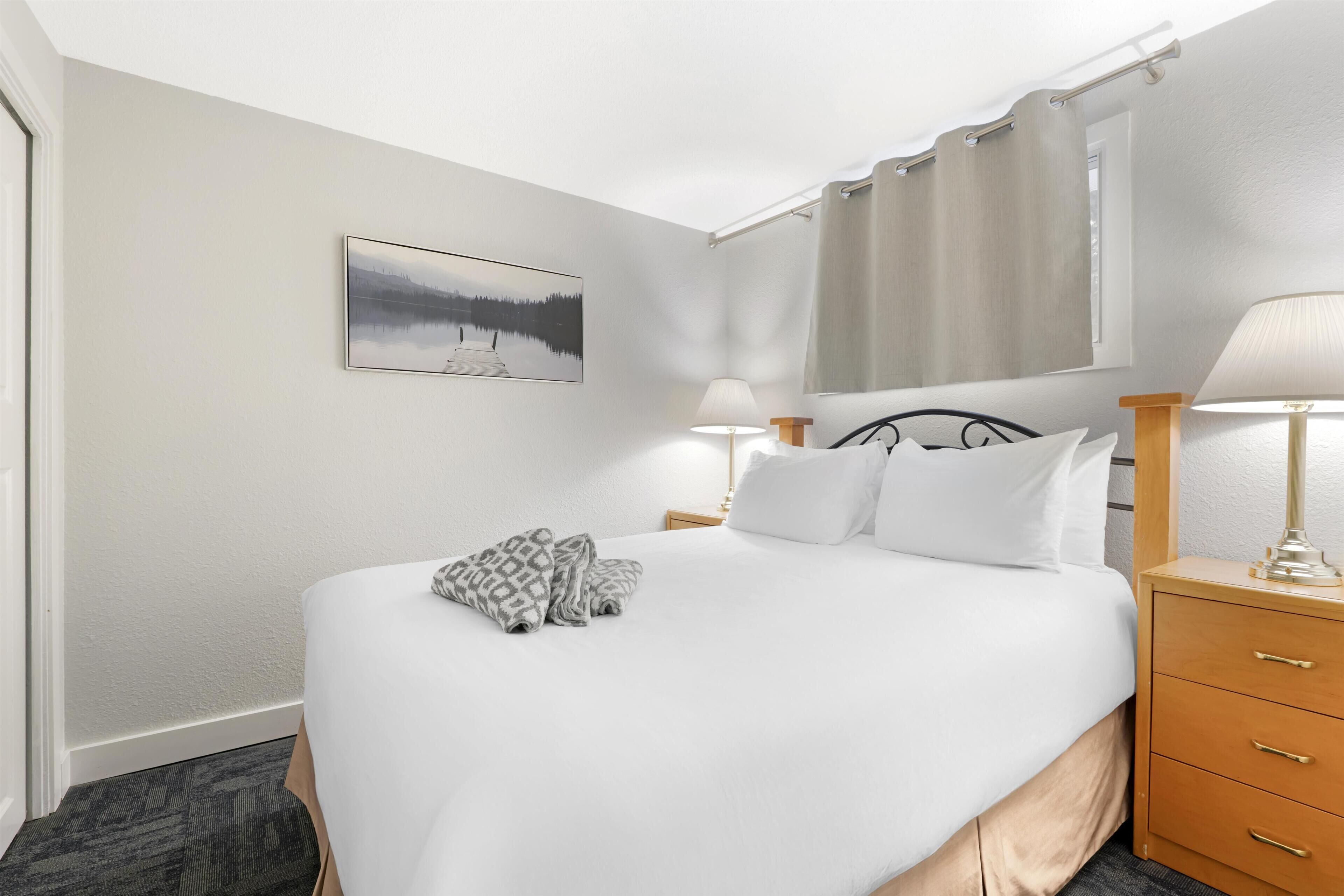 A hotel room with a bed, two bedside lamps, a wooden nightstand, a framed picture on the wall, and a window with curtains.