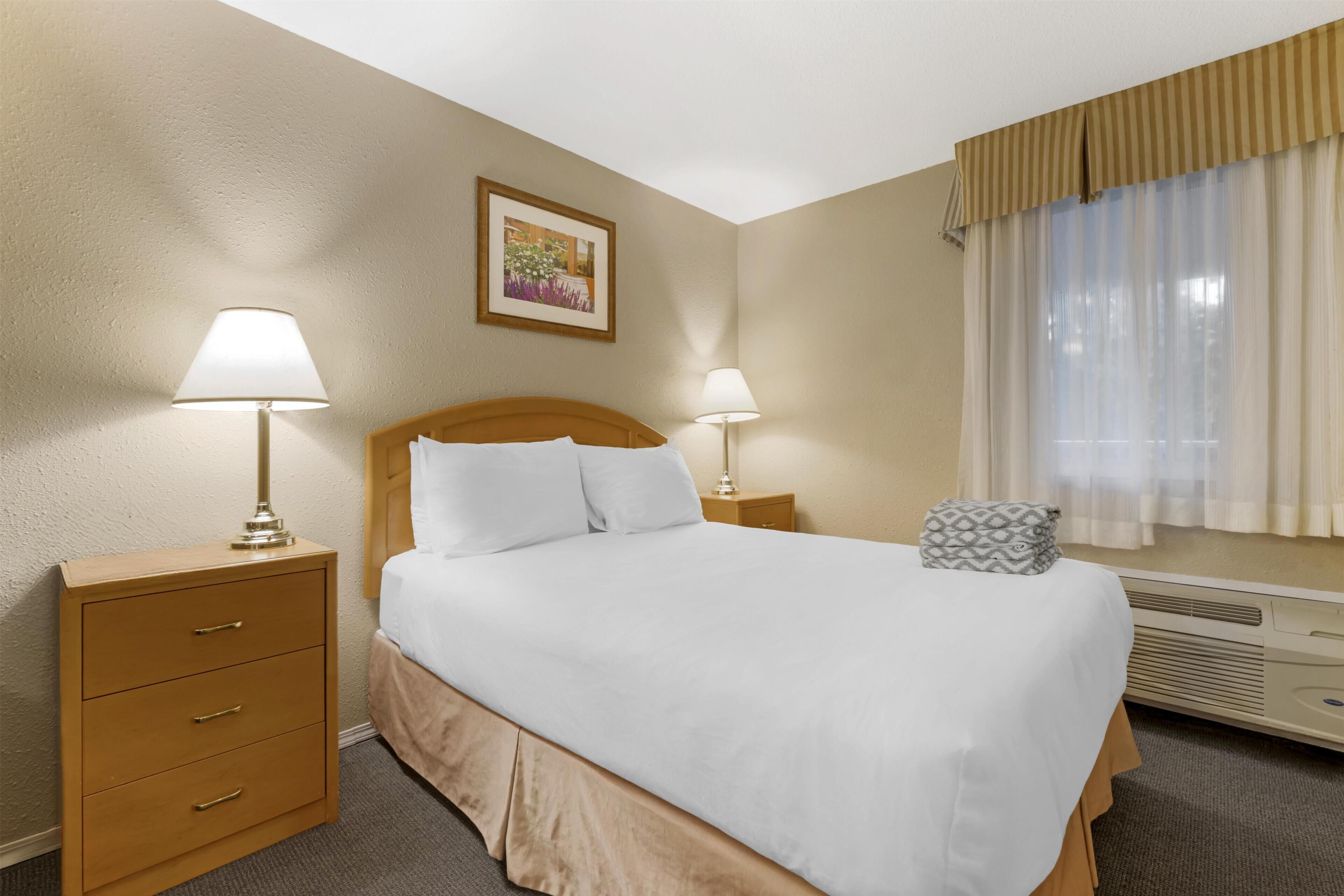 A hotel room with a bed, two bedside tables with lamps, a framed picture on the wall, and a window with curtains.