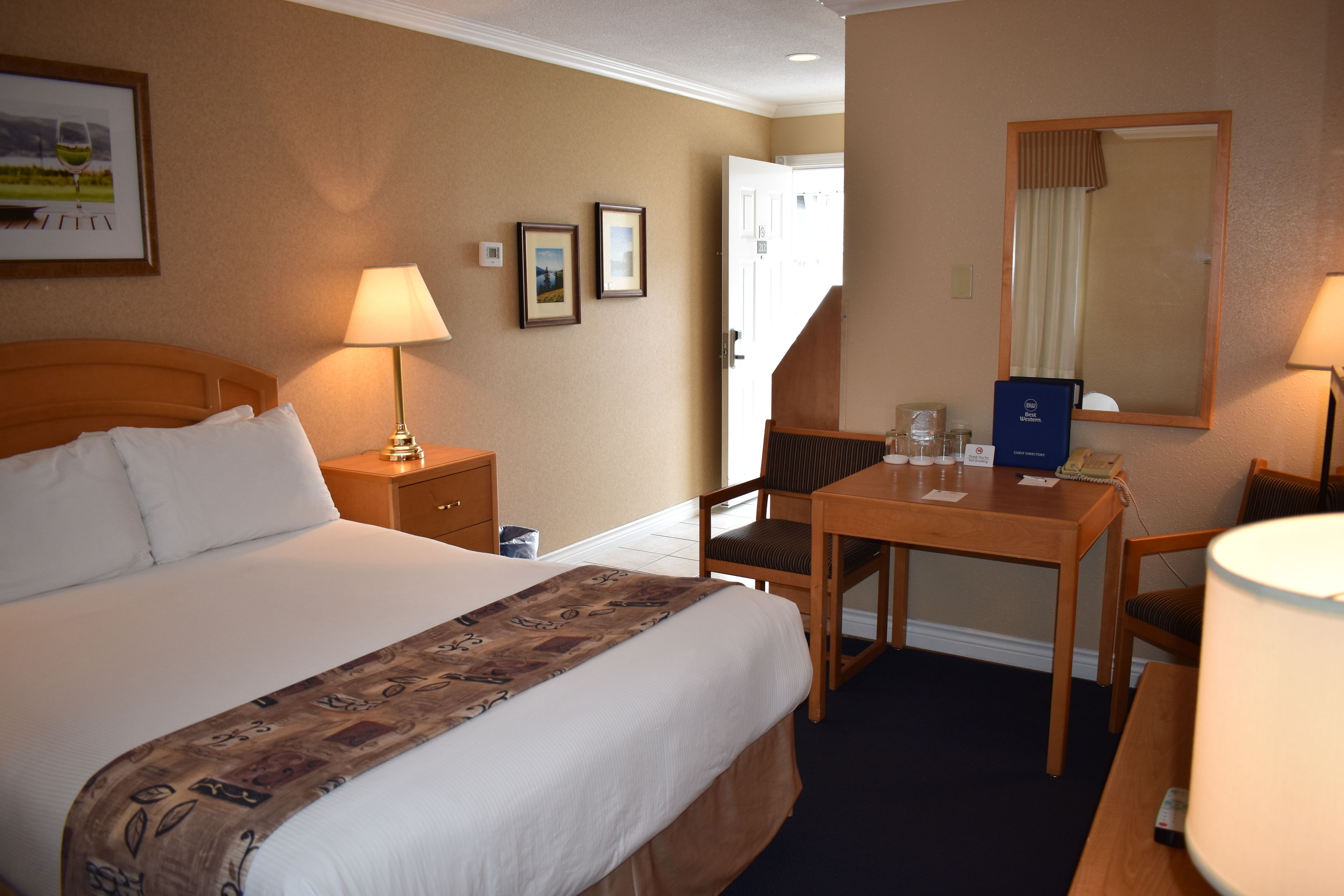 A hotel room with a large bed, a desk, a chair, a lamp, a mirror, and framed pictures on the wall.