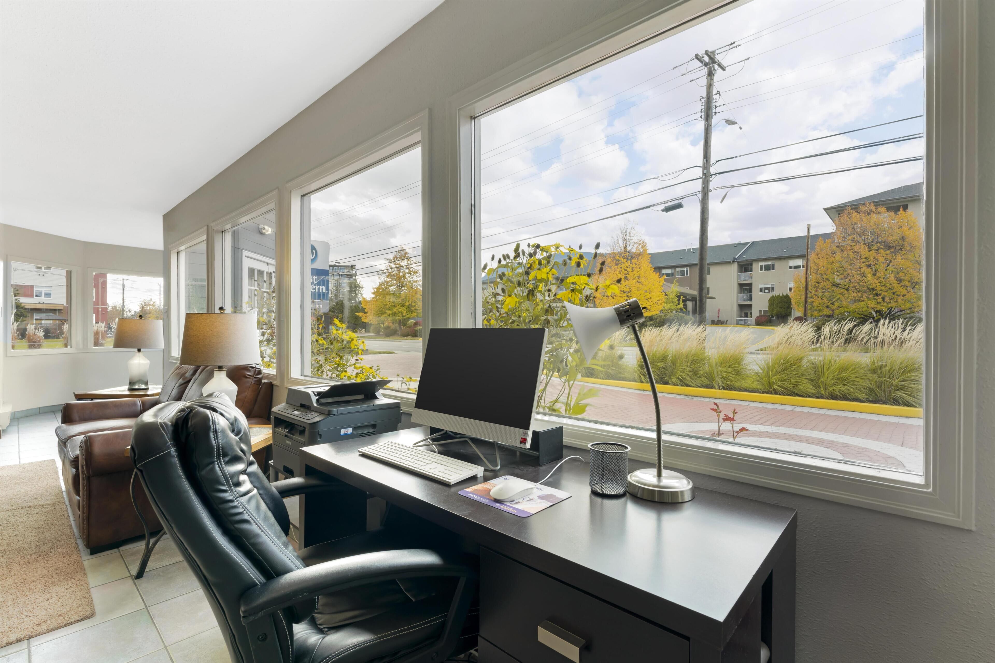 A modern office with a desk, computer, chair, lamp, and large windows offering a view of buildings and trees.