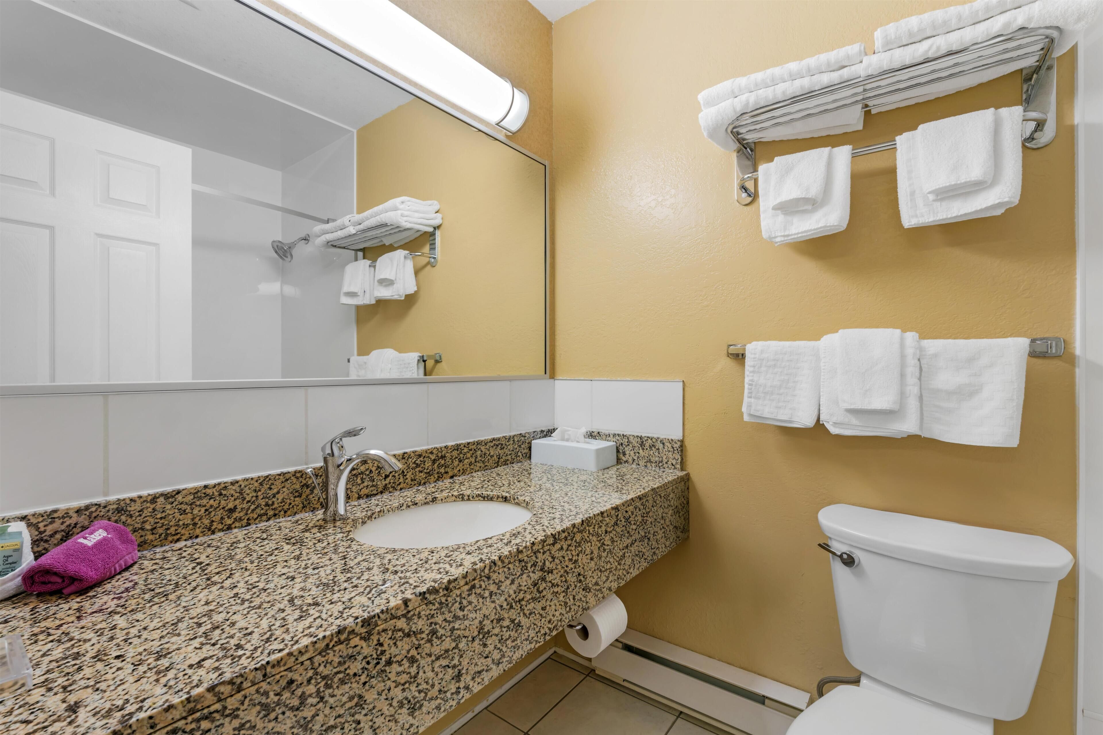 A bathroom with a granite countertop sink, a mirror, a toilet, and a towel rack with folded towels.