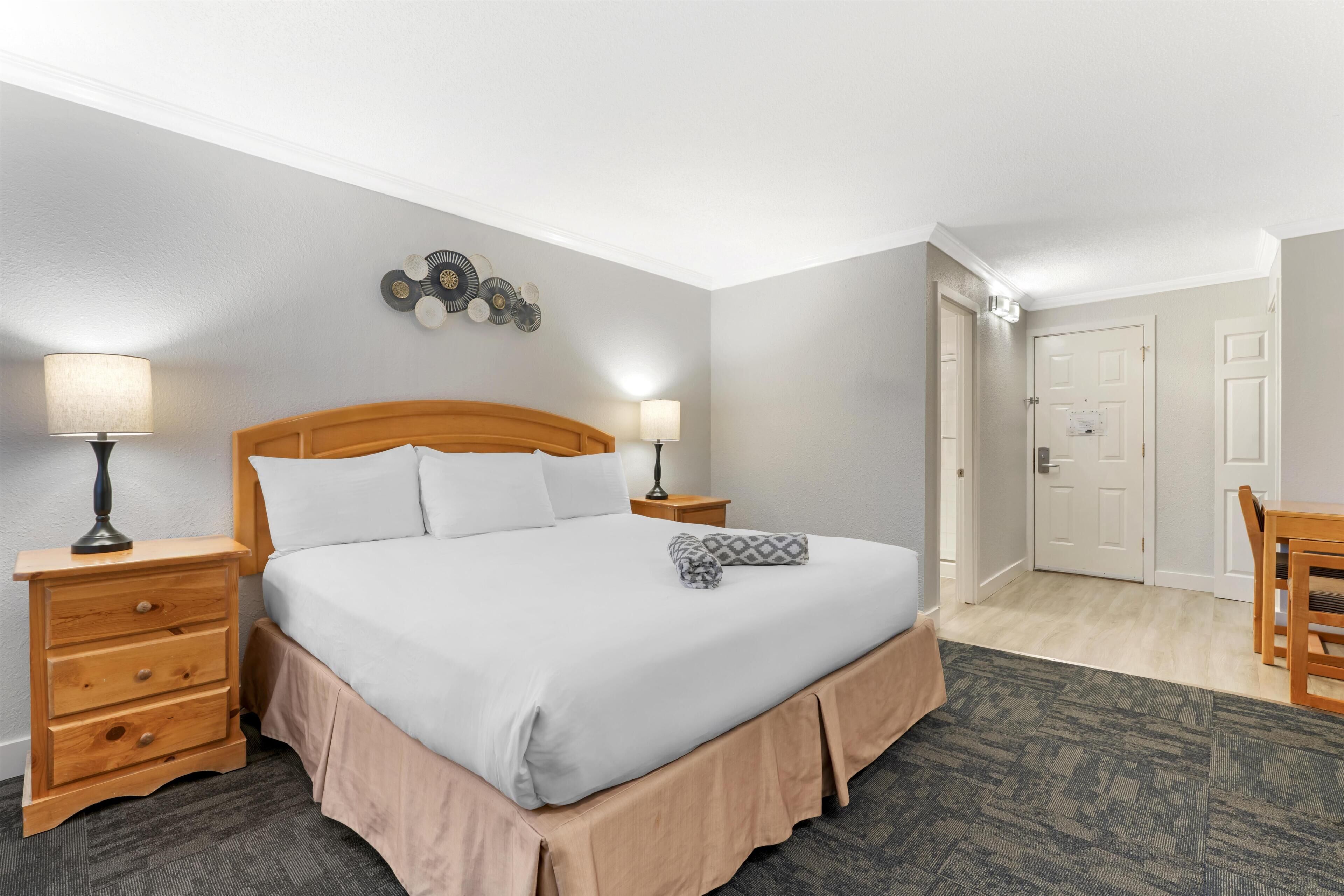 A hotel room with a bed, two bedside tables with lamps, a wall clock, and a door leading to another room.