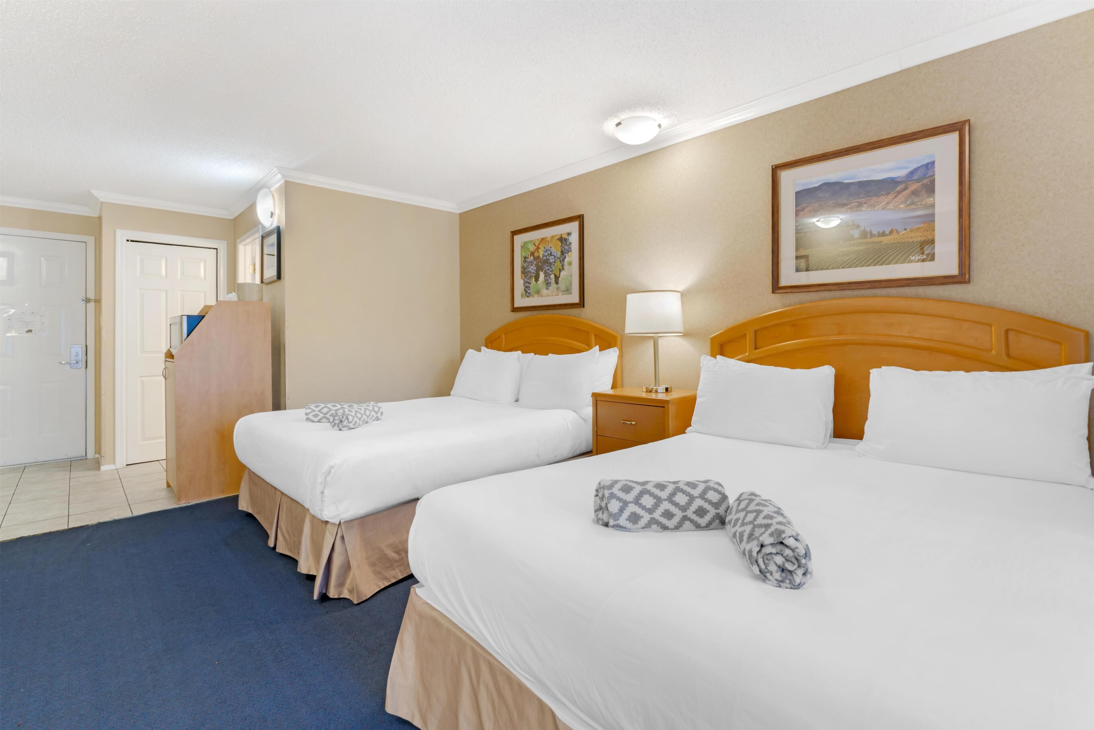 A hotel room with two beds, a desk, a lamp, and a painting on the wall.