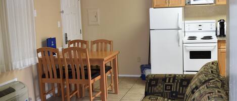 Un espacio habitable compacto con kitchenette, una zona de comedor con mesa y sillas, un sofá estampado y un ventilador de techo.