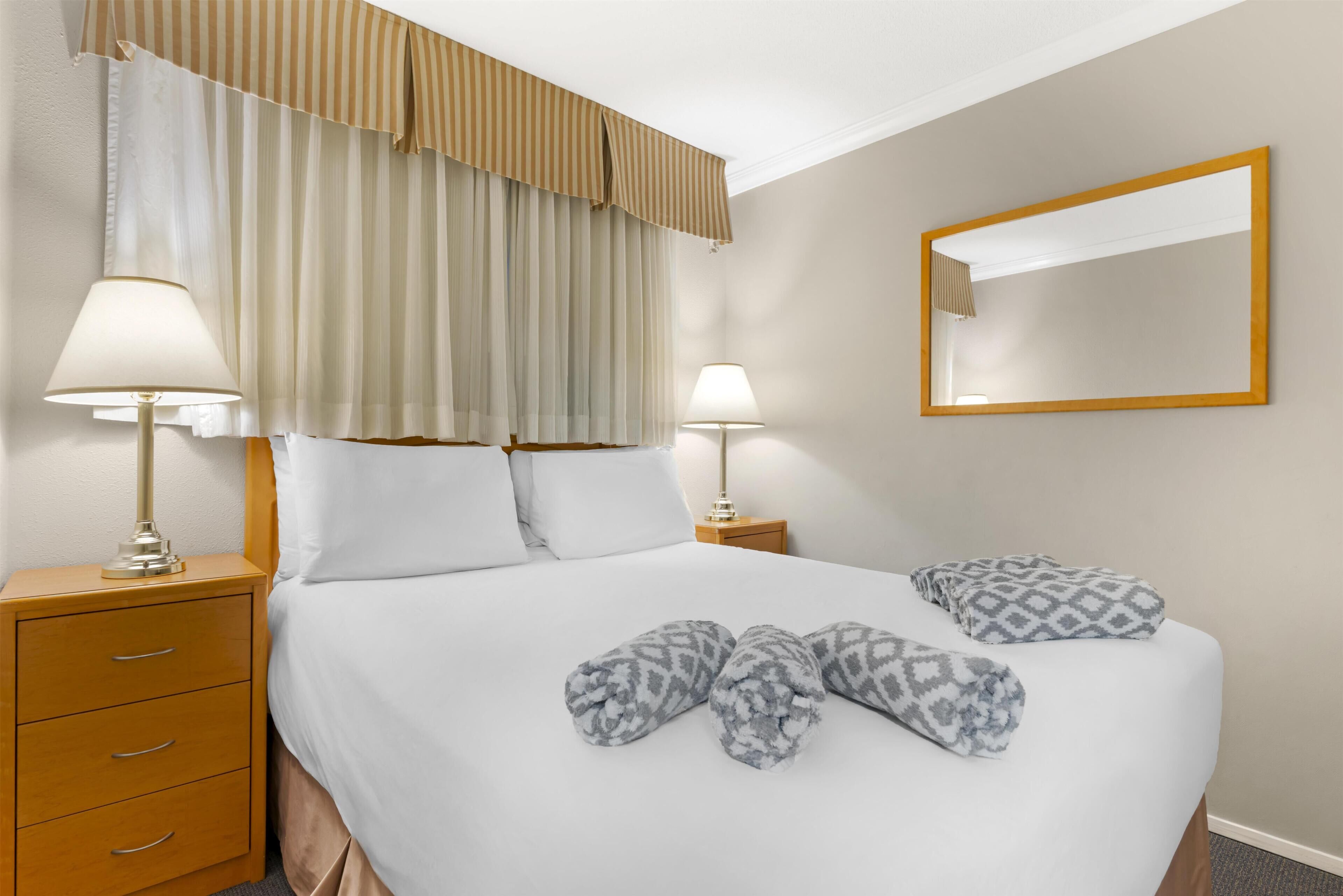 A neatly made bed with patterned pillows, two bedside lamps, a wooden nightstand with drawers, and a large mirror on the wall.
